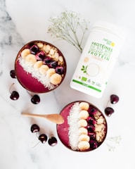 Two smoothie bowls filled with a pinkish berry blend are topped with sliced bananas, cherries, almond slices, and shredded coconut. A wooden spoon lies beside the bowls. Scattered cherries and a container of organic protein powder add to the composition on a marble surface. Some delicate white flowers are also incorporated into the arrangement.