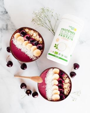 Two smoothie bowls filled with a pinkish berry blend are topped with sliced bananas, cherries, almond slices, and shredded coconut. A wooden spoon lies beside the bowls. Scattered cherries and a container of organic protein powder add to the composition on a marble surface. Some delicate white flowers are also incorporated into the arrangement.