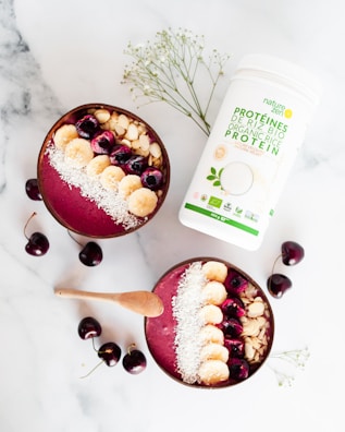 Two smoothie bowls filled with a pinkish berry blend are topped with sliced bananas, cherries, almond slices, and shredded coconut. A wooden spoon lies beside the bowls. Scattered cherries and a container of organic protein powder add to the composition on a marble surface. Some delicate white flowers are also incorporated into the arrangement.