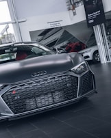A sleek, dark-colored Audi sports car is prominently displayed in a showroom, with focus on its front grille and logo. The background includes signage and other cars, enhancing a luxury and modern feel.