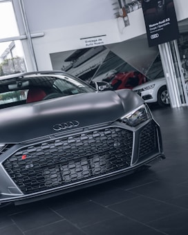A sleek, dark-colored Audi sports car is prominently displayed in a showroom, with focus on its front grille and logo. The background includes signage and other cars, enhancing a luxury and modern feel.