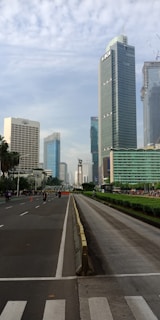 A wide street lined with tall modern skyscrapers under a cloudy sky. The road has minimal traffic with a few motorcyclists and is bordered by green hedges. The buildings have reflective glass facades with one prominent building displaying 'BCA' on its side.