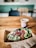A wooden table holds a cardboard tray with an open-faced sandwich topped with a variety of fresh greens, seeds, avocado slices, pomegranate seeds, and microgreens. Beside it is a disposable coffee cup with a lid and a stirring straw. The background shows a seating area with blurred chairs and a teal-green wall, providing a cozy cafe setting.