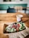 A wooden table holds a cardboard tray with an open-faced sandwich topped with a variety of fresh greens, seeds, avocado slices, pomegranate seeds, and microgreens. Beside it is a disposable coffee cup with a lid and a stirring straw. The background shows a seating area with blurred chairs and a teal-green wall, providing a cozy cafe setting.