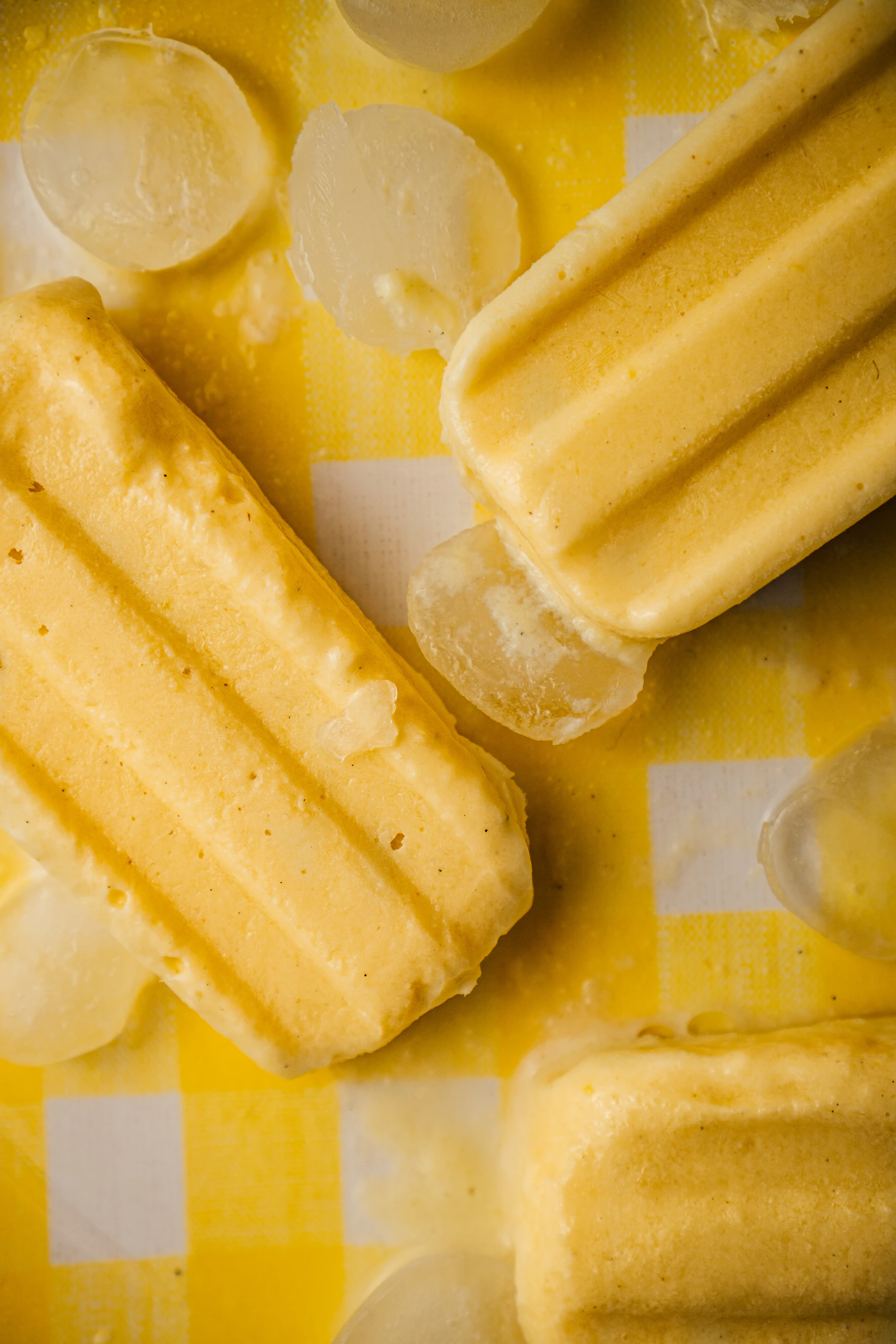 Close-up of creamy pale yellow pops lying on a yellow gingham cloth among melting ice
