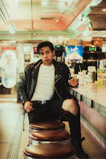A person wearing a leather jacket and jeans sits on a bar stool in a retro-style diner. The diner features a tiled floor, neon lights, and a counter with a milkshake and a menu display. Vintage decor and soft lighting enhance the nostalgic atmosphere.