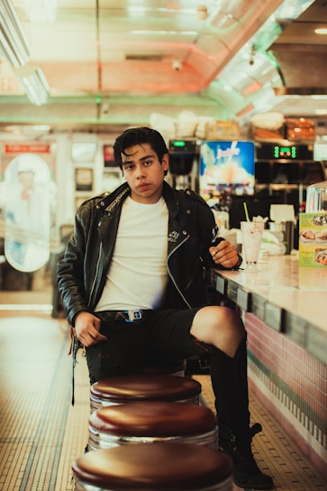 A person wearing a leather jacket and jeans sits on a bar stool in a retro-style diner. The diner features a tiled floor, neon lights, and a counter with a milkshake and a menu display. Vintage decor and soft lighting enhance the nostalgic atmosphere.