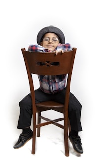 A young child sitting backwards on a wooden chair, wearing a plaid shirt, dark pants, and a flat cap. The child is smiling, with their arms resting on the back of the chair, and is wearing round glasses.