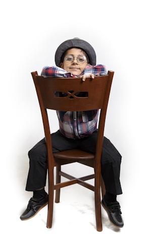 A young child sitting backwards on a wooden chair, wearing a plaid shirt, dark pants, and a flat cap. The child is smiling, with their arms resting on the back of the chair, and is wearing round glasses.