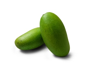 Two green mangoes are positioned against a plain white background, one leaning slightly against the other, both showing a smooth and vibrant texture.