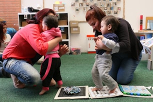 Two adults kneel on a green carpeted floor, each assisting a toddler in standing or walking. The children are exploring textured surfaces made of various materials. In the background, shelves contain bins and a wall displays photos.