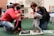 Two adults kneel on a green carpeted floor, each assisting a toddler in standing or walking. The children are exploring textured surfaces made of various materials. In the background, shelves contain bins and a wall displays photos.