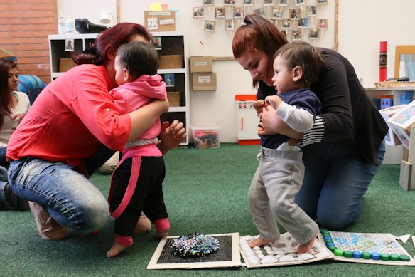 Two adults kneel on a green carpeted floor, each assisting a toddler in standing or walking. The children are exploring textured surfaces made of various materials. In the background, shelves contain bins and a wall displays photos.