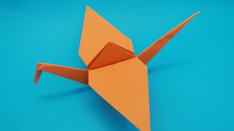 An orange origami crane is placed against a bright blue background. The crane features sharp, symmetrical folds with wings stretched out and a pointed tail. The simplicity and elegance of the origami design are highlighted by the contrast between the vivid orange paper and the vibrant blue backdrop.