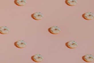 A pattern of evenly spaced donuts with white icing and multicolored sprinkles on a light pink background. The donuts cast small shadows, giving the image a sense of depth.