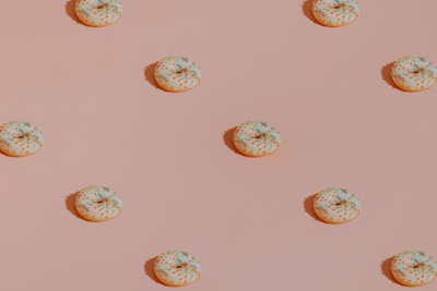A pattern of evenly spaced donuts with white icing and multicolored sprinkles on a light pink background. The donuts cast small shadows, giving the image a sense of depth.