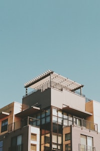 A modern multi-story building with a rooftop pergola against a clear blue sky. The structure features large glass windows, metal railings, and a combination of light and dark colored exterior surfaces.