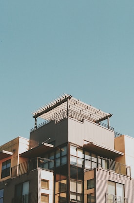 A modern multi-story building with a rooftop pergola against a clear blue sky. The structure features large glass windows, metal railings, and a combination of light and dark colored exterior surfaces.