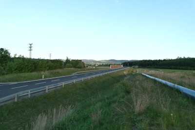 A rural road stretches towards the horizon, bordered by grassy fields and a parallel pipeline. In the background, rolling hills and a distant forest line the horizon under a clear blue sky.