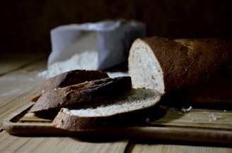A loaf of bread partially sliced sits on a wooden cutting board with a bag of flour in the background. The lighting creates a warm, rustic atmosphere.