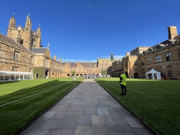 A historic university courtyard with Gothic-style architecture, surrounded by well-maintained green lawns. The scene includes a pathway leading to an ornate building in the background, with people walking and gathering. A person in a high-visibility vest stands on the edge of the lawn, beside a white tent setup.