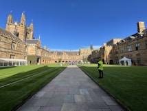 A historic university courtyard with Gothic-style architecture, surrounded by well-maintained green lawns. The scene includes a pathway leading to an ornate building in the background, with people walking and gathering. A person in a high-visibility vest stands on the edge of the lawn, beside a white tent setup.