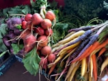 A vibrant assortment of fresh vegetables, including bunches of beets and multi-colored carrots, resting on a flat surface. The beets are deep red and purple, with lush green leaves still attached. The carrots vary in color, featuring shades of purple, orange, yellow, and white. In the background, a dense layer of green leafy tops adds to the fresh, natural aesthetic.