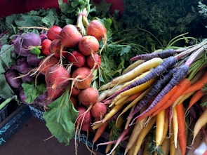 A vibrant assortment of fresh vegetables, including bunches of beets and multi-colored carrots, resting on a flat surface. The beets are deep red and purple, with lush green leaves still attached. The carrots vary in color, featuring shades of purple, orange, yellow, and white. In the background, a dense layer of green leafy tops adds to the fresh, natural aesthetic.