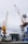 A large boat is positioned in a shipyard, flanked by two towering cranes. The boat is elevated off the ground, suggesting it is undergoing maintenance or construction. A small white truck is parked in front of the boat, and various industrial structures and equipment surround the area. The sky is overcast.