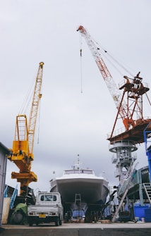 A large boat is positioned in a shipyard, flanked by two towering cranes. The boat is elevated off the ground, suggesting it is undergoing maintenance or construction. A small white truck is parked in front of the boat, and various industrial structures and equipment surround the area. The sky is overcast.