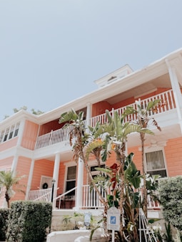 A pastel-colored building with a peach facade and white railings, complemented by tropical plants and a reserved parking sign in the foreground. The sky is clear and blue, creating a bright and inviting atmosphere.