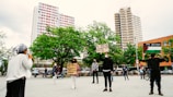 A group of people gathers in a public square, with a backdrop of tall buildings and green trees. Individuals hold signs with messages supporting Palestine, and one person speaks into a microphone. The participants maintain social distancing and wear masks, indicating a socially-aware demonstration.