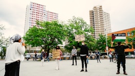 A group of people gathers in a public square, with a backdrop of tall buildings and green trees. Individuals hold signs with messages supporting Palestine, and one person speaks into a microphone. The participants maintain social distancing and wear masks, indicating a socially-aware demonstration.