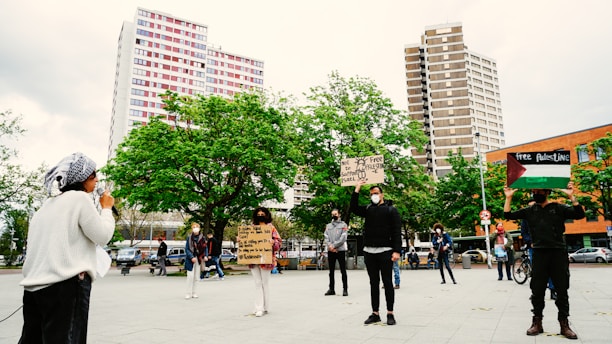 A group of people gathers in a public square, with a backdrop of tall buildings and green trees. Individuals hold signs with messages supporting Palestine, and one person speaks into a microphone. The participants maintain social distancing and wear masks, indicating a socially-aware demonstration.