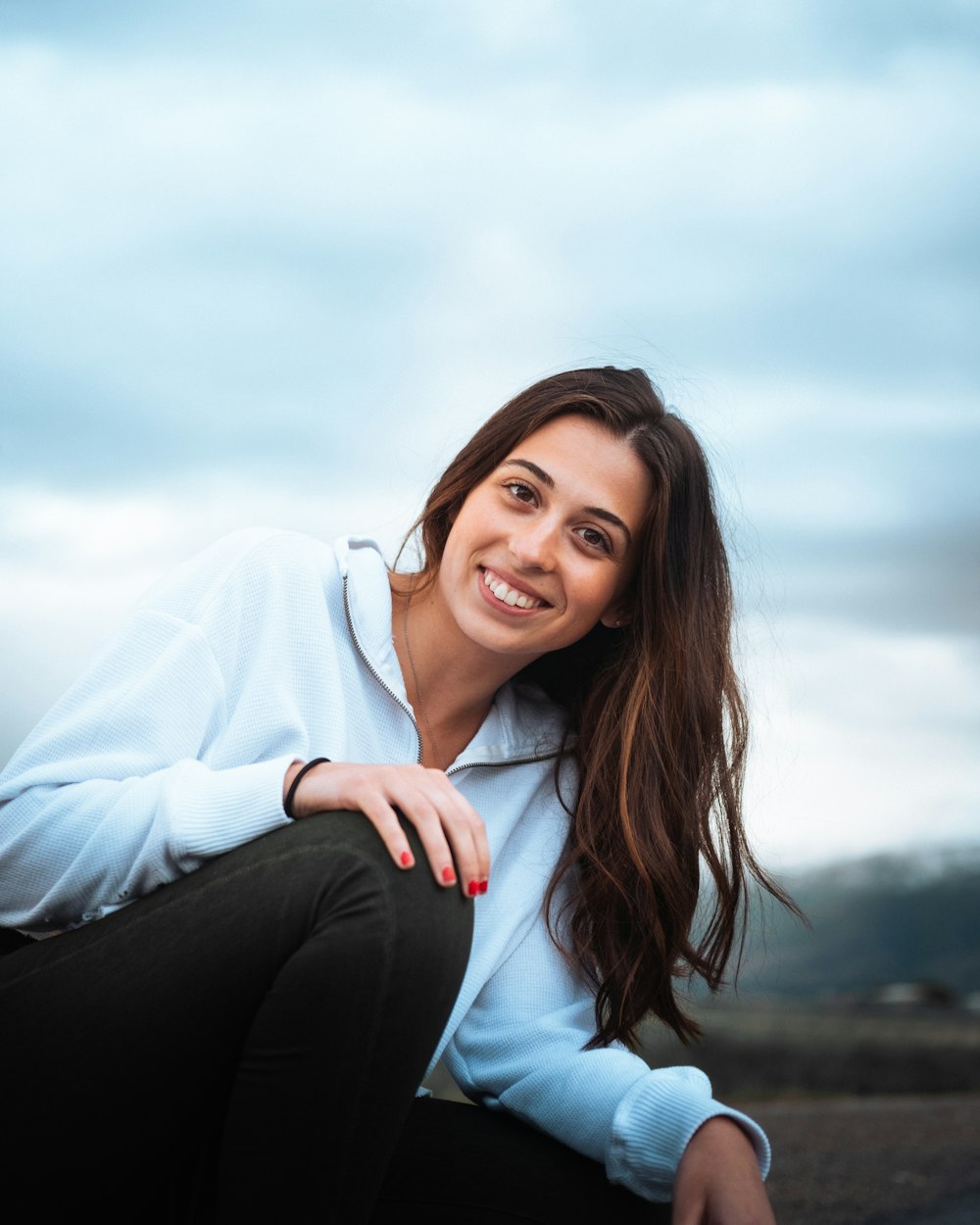 woman in white long sleeve shirt and black pants sitting on gray concrete pavement during daytime