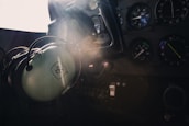 A cockpit interior with a close-up view of a control panel and dials. A pair of aviation headphones is hanging prominently, with sunlight streaming through the window creating a dramatic highlight.