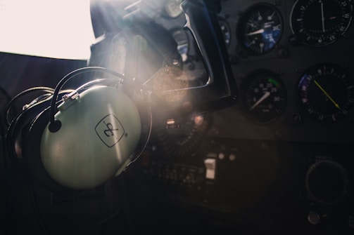 A cockpit interior with a close-up view of a control panel and dials. A pair of aviation headphones is hanging prominently, with sunlight streaming through the window creating a dramatic highlight.