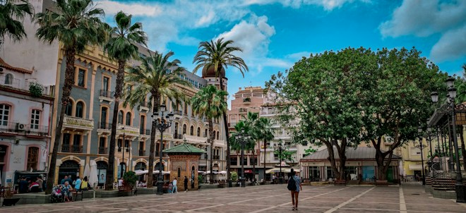 A vibrant city square features historical buildings with ornate architecture, lined with palm trees. The open area is bordered by a mix of classical and modern structures, with people strolling and engaging in activities. The sky is partly cloudy, adding depth to the scene.