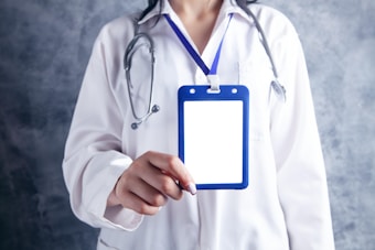A person wearing a white lab coat is holding a blank ID badge with a blue lanyard. A stethoscope is draped around the neck, suggesting a medical professional. The background is a textured gray.