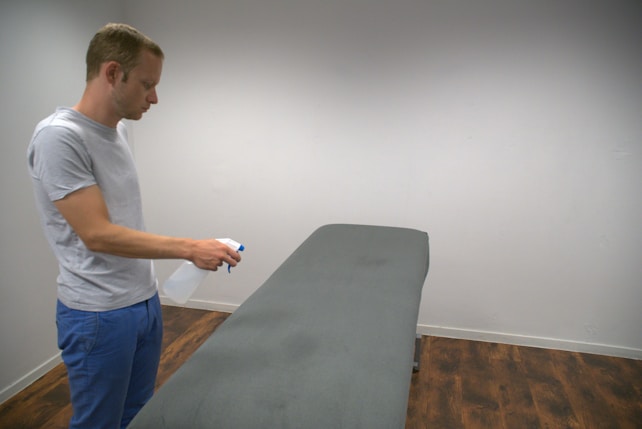 A person in casual clothing holds a spray bottle while standing next to a massage table. The table is covered with a dark gray sheet, and the setting is a simply furnished room with wooden flooring and white walls.