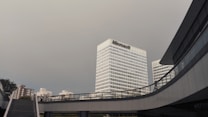 A modern urban scene featuring a large office building with the Microsoft logo prominently displayed. The building is surrounded by other tall structures, along with sky bridges and a staircase in the foreground, under a cloudy sky.