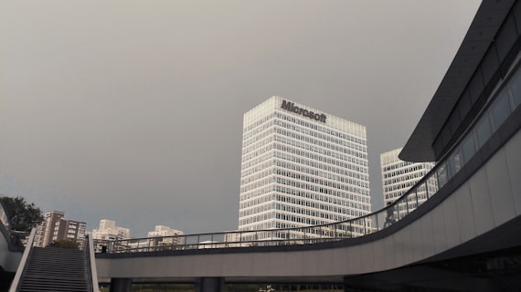 A modern urban scene featuring a large office building with the Microsoft logo prominently displayed. The building is surrounded by other tall structures, along with sky bridges and a staircase in the foreground, under a cloudy sky.