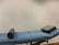 A massage or examination table with a blue cover is centered in the image. On the table lies a cylindrical bolster and a square cushion, both in a dark grey material. The background features a plain, light-colored wall.