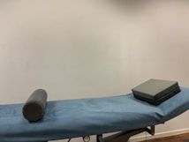 A massage or examination table with a blue cover is centered in the image. On the table lies a cylindrical bolster and a square cushion, both in a dark grey material. The background features a plain, light-colored wall.