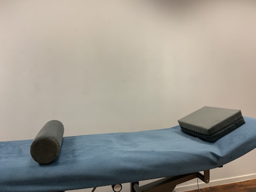 A massage or examination table with a blue cover is centered in the image. On the table lies a cylindrical bolster and a square cushion, both in a dark grey material. The background features a plain, light-colored wall.