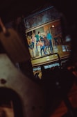 A vibrant illustration featuring several characters in dynamic poses, possibly engaged in a confrontation, set in an urban environment with vintage attire. The scene is visible through a dark, blurred foreground which frames the image.