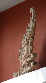 A decorative stone sculpture with intricate carvings, including floral and circular patterns, situated against a textured, rust-colored wall. The sculpture features elaborate curves and layered details, resembling a stylized wave or crest.