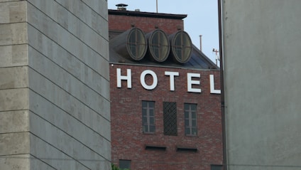 A building with a brick exterior prominently displays the word 'HOTEL' in large white letters on its facade. The building features round, circular structures on the roof and is partially obscured by two other buildings on either side.