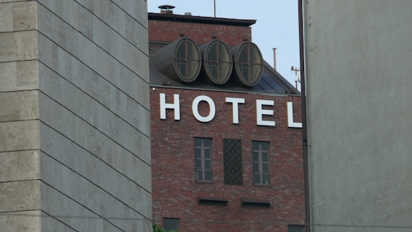 A building with a brick exterior prominently displays the word 'HOTEL' in large white letters on its facade. The building features round, circular structures on the roof and is partially obscured by two other buildings on either side.