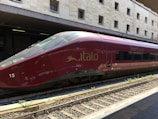 A sleek red high-speed train is stationed on railway tracks at a modern train station, with a beige stone building in the background. The text 'Italo' and 'Smart Cinema' are visible on the side of the train, indicating its branding.
